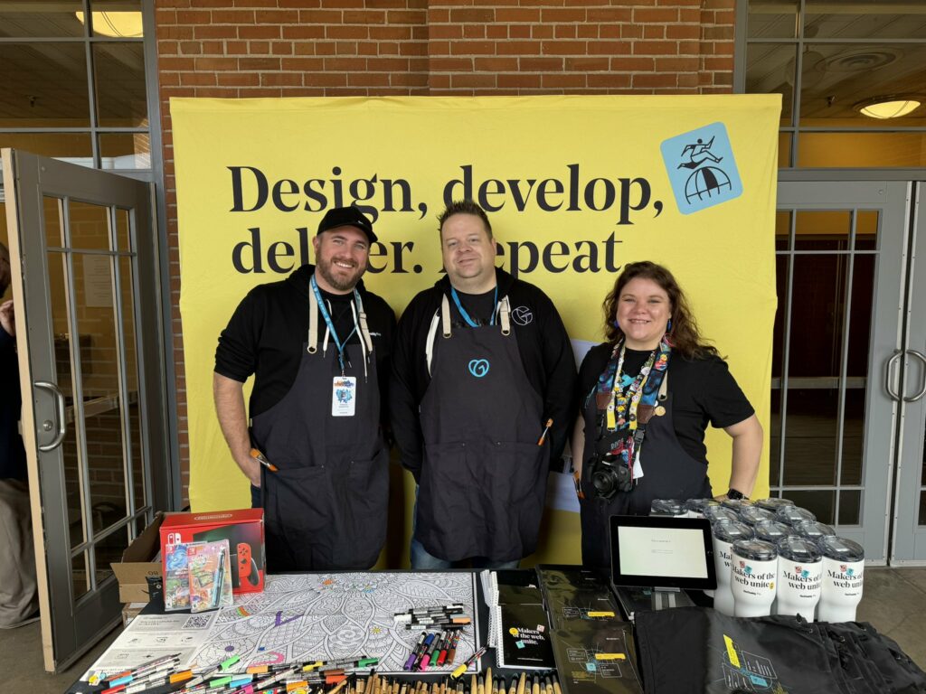 Three individuals standing in front of a yellow banner with the text “Design, develop, deliver, repeat” and a logo of a carousel horse. To their front is a table with various items including t-shirts, cups, a coloring sheet with markers, a Nintendo Switch box, and a tablet displaying a message. They wear black aprons and lanyards with ID badges.