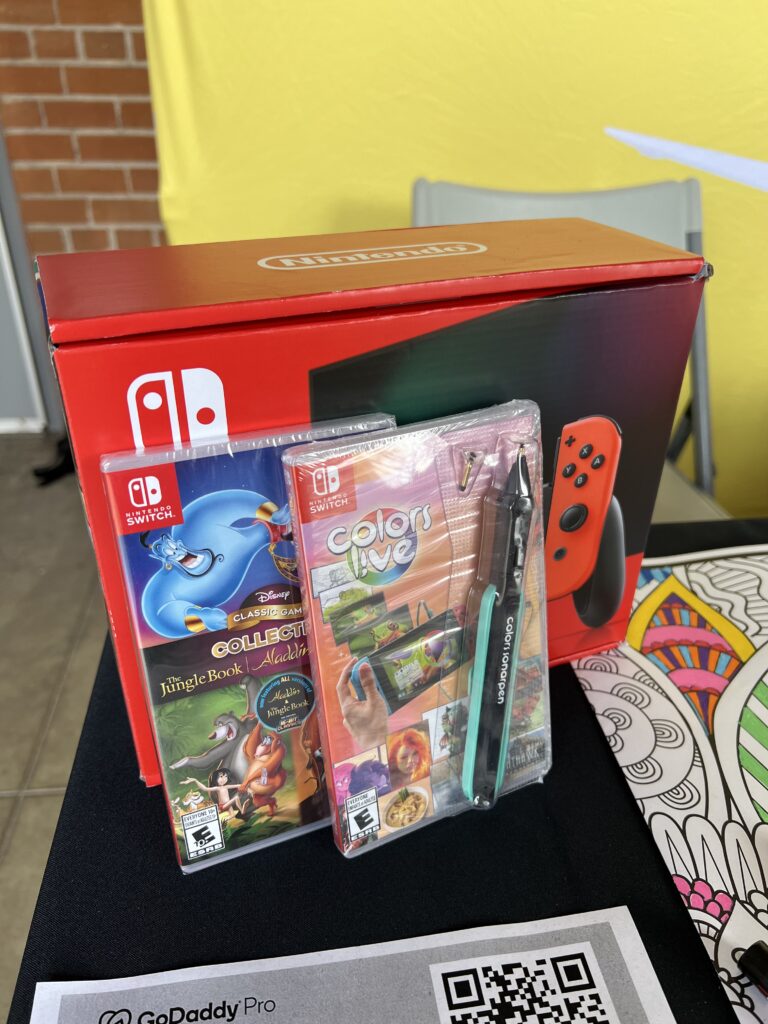 A Nintendo Switch box with its content partially displayed, including game cases for ‘Disney Classic Games: Aladdin and The Lion King’ and ‘Colors Live,’ along with a color pen accessory, placed on a black surface with a QR code and against a backdrop featuring a yellow wall and a part of a gray chair.